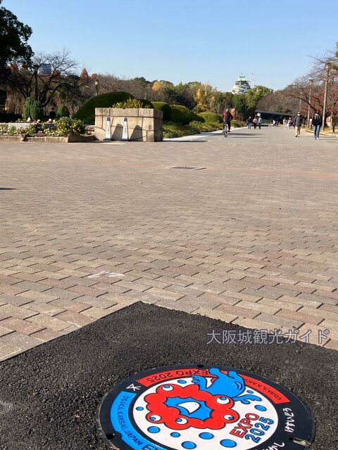 大阪城公園にあるミャクミャク様の「大阪・関西万博デザインマンホールふた」。奥に大阪城天守閣とスターバックスコーヒーが見える